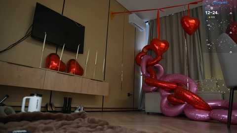 Video of a cozy, modern living room with a flat-screen TV mounted on a beige wall, red balloons, and a pink inflatable snake on the floor, creating a playful, festive atmosphere.