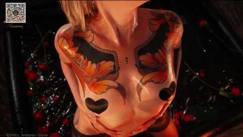Video of a topless woman with tattoos of orange fish and black hearts on her chest, lying in a dark, wet environment surrounded by red flowers.