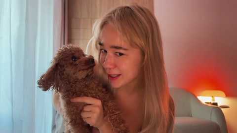Video of a young blonde woman with long hair, fair skin, and a small, curly brown dog. She wears a white top, smiles, and holds the dog. Background features a soft, warm light, a lamp, and light-colored walls.