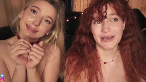 Media: A video of two topless women with fair skin and long hair, one blonde and the other red, smiling and holding hands, in a dimly lit room with string lights.