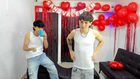 Video of two teenage boys in white tank tops, blue jeans, and blue face masks, standing in a room with red heart decorations, balloons, and a \"SANDBOX\" banner.