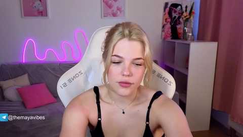 Video of a young woman with fair skin and blonde hair, wearing a black camisole, seated in a gray chair with a pink neon light, surrounded by floral decor and a desk with art supplies.
