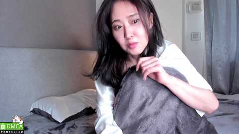Media: Video of a young Asian woman with long black hair, fair skin, and medium build, wearing a white shirt and gray blanket, sitting on a bed with white pillows and gray sheets in a modern, softly lit room.