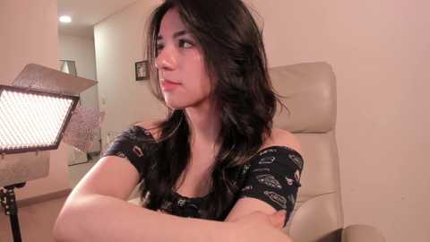 Media: Video of a young woman with long, wavy black hair, wearing a black top with car logos, sitting in a beige office chair, looking thoughtful, in a dimly lit room with a metal lamp and a blurred background.