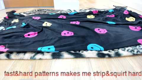 Video of a black fabric with colorful, cartoonish cat faces and ears, draped on a leopard-print blanket on a light wooden floor, with text overlay in red and orange.