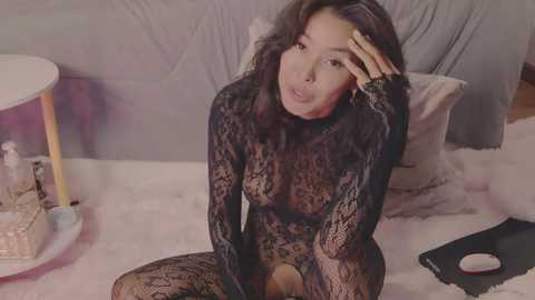 Video of an Asian woman with long black hair, wearing a sheer black lace bodysuit, sitting on a bed in a pink room, touching her forehead, with a mouse on the floor.