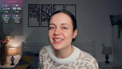 Video of a smiling woman with light skin, brown hair, wearing a leopard print sweater, in a modern bedroom with white walls, black tree wall art, and a lit lamp.
