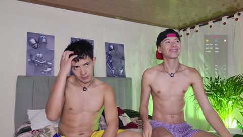 Video of two shirtless, slender Asian men in boxer shorts, sitting on a bed with a green headboard. They wear black headbands and black necklaces. Background shows posters, green curtains, and potted plants.