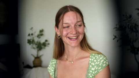 Video of a smiling, fair-skinned woman with long, straight brown hair, wearing a green floral dress and gold hoop earrings, in a softly lit, dimly lit room with a potted plant in the background.