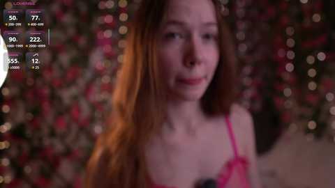 Video of a young woman with long, auburn hair, wearing a pink lace bra. Background is blurred with festive lights.