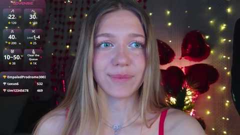 Video of a young, light-skinned woman with long, straight blonde hair, blue eyes, and a slight smile. She wears a red spaghetti strap top, surrounded by dimly lit, red heart-shaped decorations and fairy lights in the background.