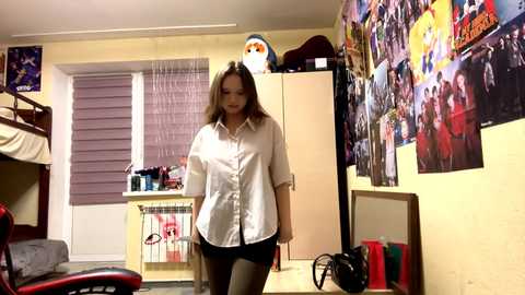 Video of a young woman in a white button-up shirt and black skirt, standing in a cluttered, colorful college dorm room with posters, a bunk bed, and a window with blinds.
