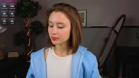 Video of a young Caucasian woman with straight, shoulder-length brown hair, wearing a white tank top and light blue jacket. Background includes a green plant, wooden chair, and a framed picture.