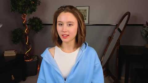 A video of a young Caucasian girl with shoulder-length brown hair, wearing a light blue hoodie over a white t-shirt, standing indoors near a green plant and a wooden chair.