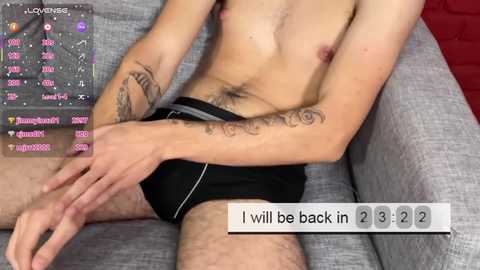 Video of a light-skinned, slender man with small breasts, wearing black underwear, sitting on a grey couch, with a text overlay reading \"I will be back in 3.12.\