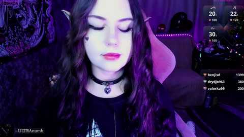 Video of a young woman with pale skin, long dark hair, and elf ears, wearing a black choker and dark clothing, against a purple-lit, dimly-lit background with a gaming setup.