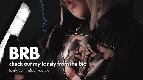 A close-up video of a woman with blonde hair, wearing a black, see-through top revealing intricate tattoo designs on her chest. She holds a flashlight. Text overlays the image, reading \"BRB out my fancy from the big\" and \"fansly.com/dice-kosmos.\