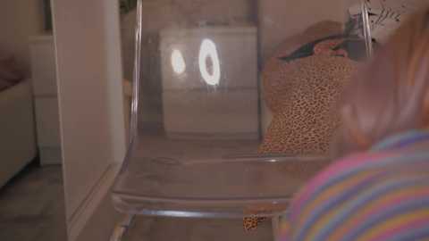 A video of a clear plastic chair with a textured brown woven bag, placed in a modern, beige-walled room with a white toilet in the background.