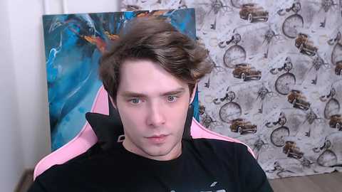 Video of a young, fair-skinned man with brown hair in a video game chair, wearing a black shirt. Behind him is a wall with a blue abstract painting and a patterned curtain featuring vintage cars.