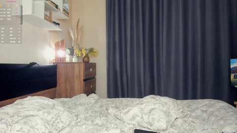 Video of a modern, cluttered bedroom with a dark gray curtain, a wooden dresser holding decorative items, a white wall-mounted shelf, and a bed with white patterned sheets.