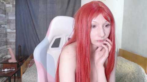 Video of a fair-skinned woman with long red hair, wearing a white and pink gaming chair, indoors with a bed, stone wall, and a wooden nightstand.