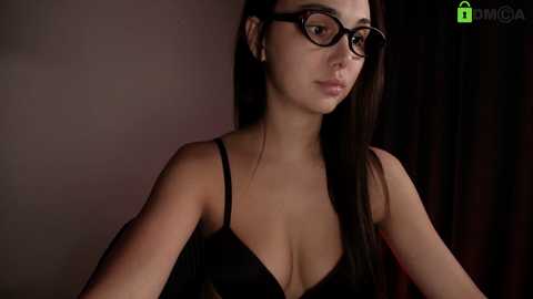 A video of a young, light-skinned woman with long, dark hair, wearing black-rimmed glasses and a black bra. She has a neutral expression, and the background is dark and dimly lit.