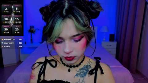 Video of a young woman with fair skin, dark hair in double buns, wearing bold red eyeshadow and black lace lingerie, featuring a floral tattoo. Background shows a dimly lit bedroom with blue lighting, and a bed.