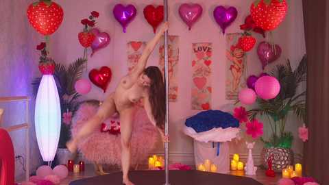 Video of a nude woman with long brown hair, dancing on a pole in a pink room decorated with heart-shaped balloons, posters, and candles.