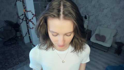 A young Caucasian woman with shoulder-length brown hair, wearing a white t-shirt and a silver necklace, stands in a dimly lit room with grey walls, a chair, and Christmas decorations.