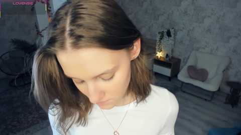 A video of a young woman with shoulder-length brown hair, wearing a white top, in a dimly lit room with floral wallpaper, a chair, and a lamp.