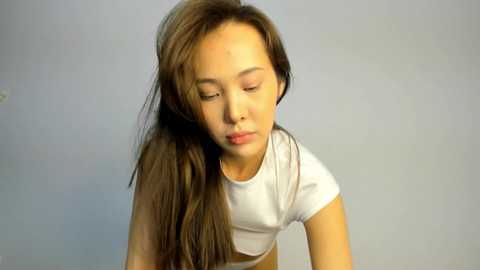 Video of a young East Asian woman with long, straight brown hair, wearing a white short-sleeved top, appearing sad and looking down. Background is plain light gray.