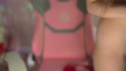 A blurred video shows a person's upper body and face in a dimly lit room, with a pink, cartoonish character in the background. The image is out of focus, making details indistinguishable.