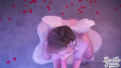 A video of a nude woman with pale skin, blonde hair, and white angel wings, kneeling on a plush white rug in a dimly lit room scattered with red rose petals.