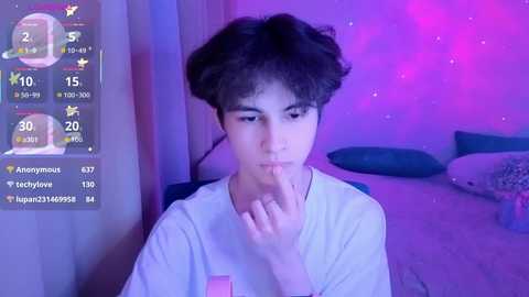 Video of a young man with curly hair, wearing a white t-shirt, sitting on a chair, touching his lips in a bedroom with purple lighting and a bed in the background.