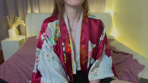 Video of a woman with light skin and brown hair, wearing a red kimono with floral patterns, sitting on a bed with a lavender sheet, in a softly lit, minimalistic bedroom.