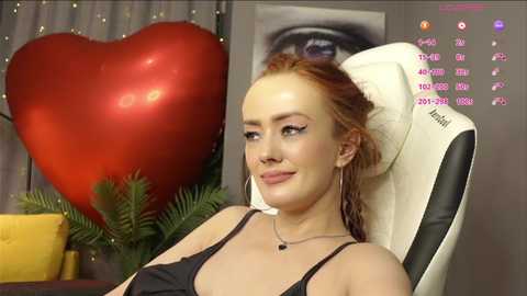 Video of a fair-skinned, red-haired woman with a slender physique, wearing a black lace bra, sitting in a white chair. Background includes a red heart-shaped cushion and a framed eye painting.