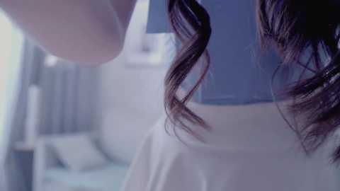 Video of a woman with long, dark brown hair, styled in loose waves. She is wearing a white shirt, with a blurred, modern, light-colored room in the background. The image has a soft, ethereal blue hue.