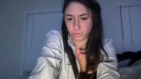 A video of a young woman with long dark hair, wearing a striped shirt, sitting on a bed with white blinds in the background.