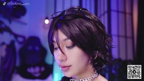 Video of an Asian woman with short, dark hair, wearing sunglasses, a silver choker, and a black top. She's in a dimly lit room with purple lighting and blurred background.