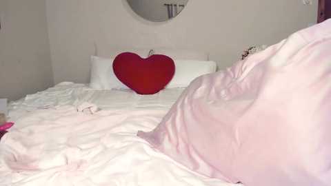 Video of a cozy, pastel-colored bedroom with a white bed, pink blanket, and a large red heart pillow, set against a soft, cream-colored wall.