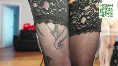Video of a person's legs wearing sheer black lace stockings with floral patterns, visible tattoos, and red rose petals scattered on a wooden floor. Background includes a black couch and a red object.