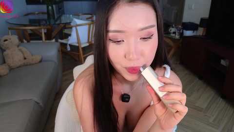 Media: A video of an East Asian woman with long black hair, wearing pink lipstick, applying eyeliner with a white pencil in a modern living room.