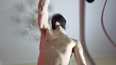 Video of a shirtless, fair-skinned man with dark hair, arms raised in a vigorous stretch, indoors with a ceiling fan and light fixture in the background.