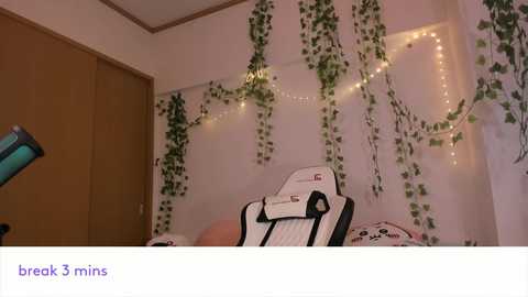 Video of a cozy bedroom with a white wall adorned with green ivy and fairy lights, featuring a black and white gaming chair with a backpack on it, and a wooden door in the background.
