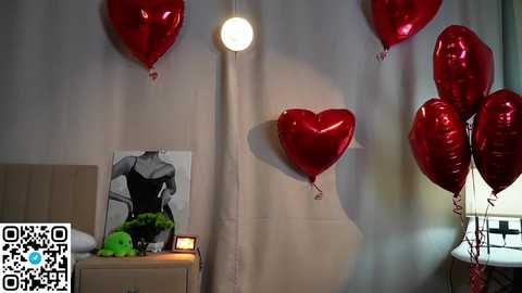 Video of a modern bedroom with a white wall, a small wooden nightstand, a black-and-white abstract painting, and six red heart-shaped balloons with a glowing white light.