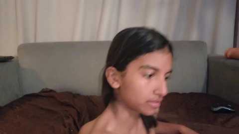 A video of a young woman with long dark hair, wearing a black top, sitting on a bed with brown sheets and a grey headboard. The background includes a beige curtain.