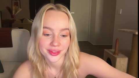 Video of a blonde woman with fair skin, wearing red lipstick, smiling, sitting in a cozy living room with a beige couch, a wooden cat scratcher, and a door in the background.