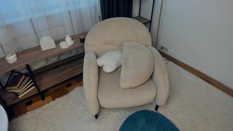 Video of a beige armchair with a white cushion, positioned in a minimalist room with a white carpet, wooden floor, and white curtains.