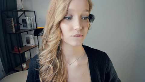 Video of a fair-skinned woman with long, wavy blonde hair, wearing black clothing, and makeup with dark eyeliner, looking contemplative indoors.