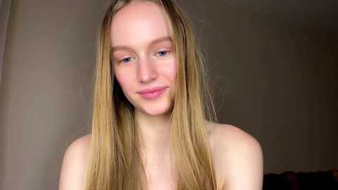 Video of a young, fair-skinned woman with long, straight blonde hair, wearing minimal makeup, and appearing serene. She is topless, set against a plain, neutral-colored background.
